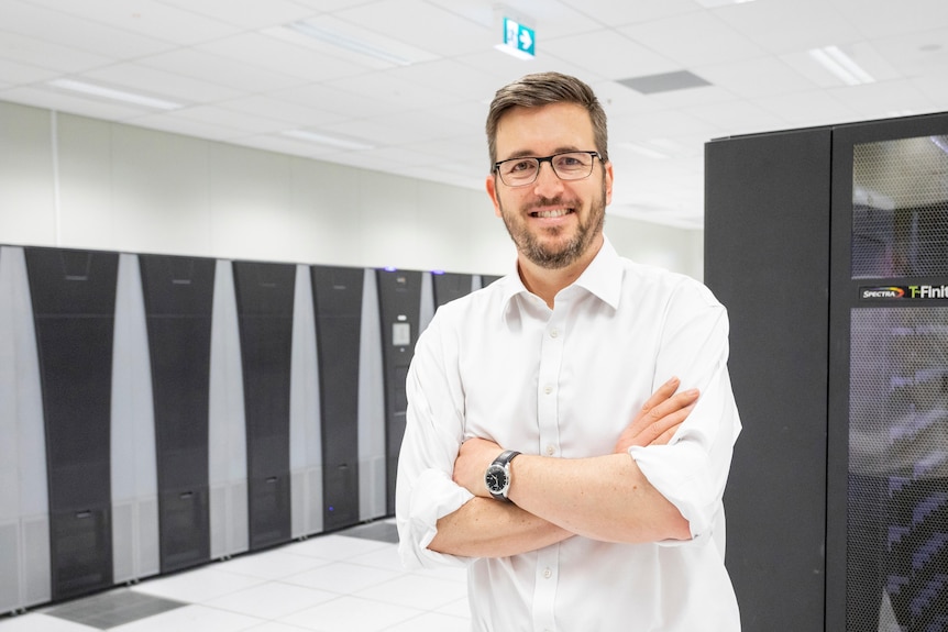 Man standing in white room in front of a supercomputer with wires and racks 