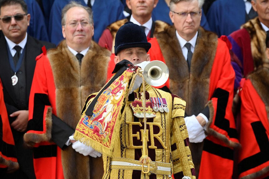 A trumpeter dressed in gold plays in front of men in red and fur robes. 