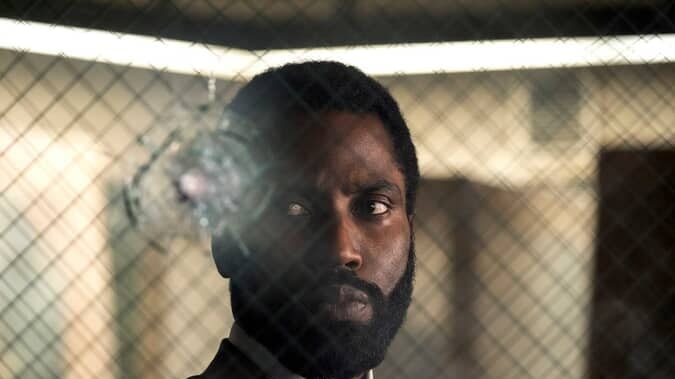 Actor John David Washington stands in a suit looking through glass that has a bullet hole in it. The picture is taken from Tenet