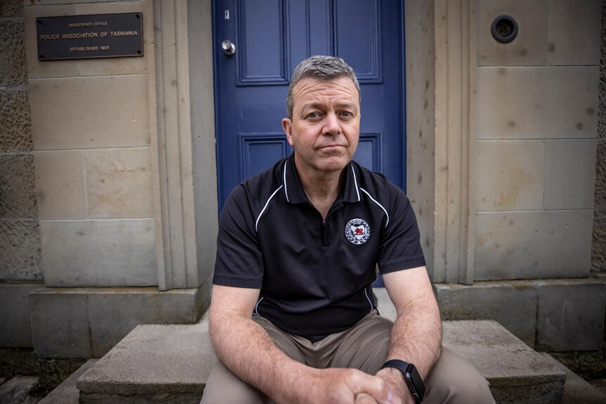 A grey-haired man sitting on steps near a blue door on a sandstone building, wearing a Police Association of Tasmania shirt