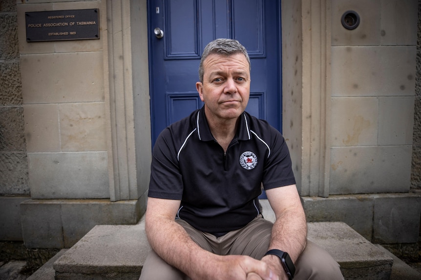 A grey-haired man sitting on steps near a blue door on a sandstone building, wearing a Police Association of Tasmania shirt