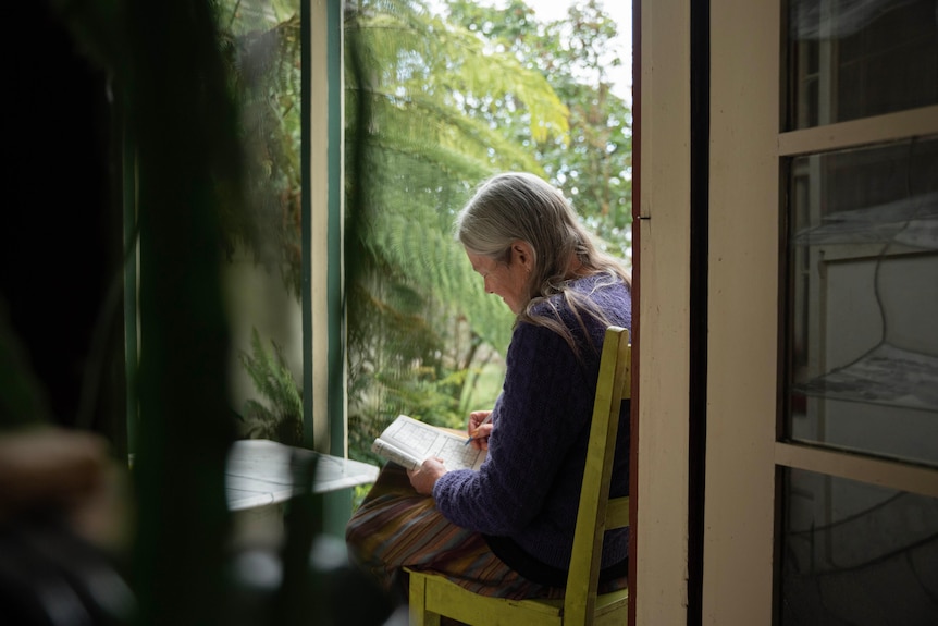 A woman sits by a window, working on a sudoku puzzle in a notebook.