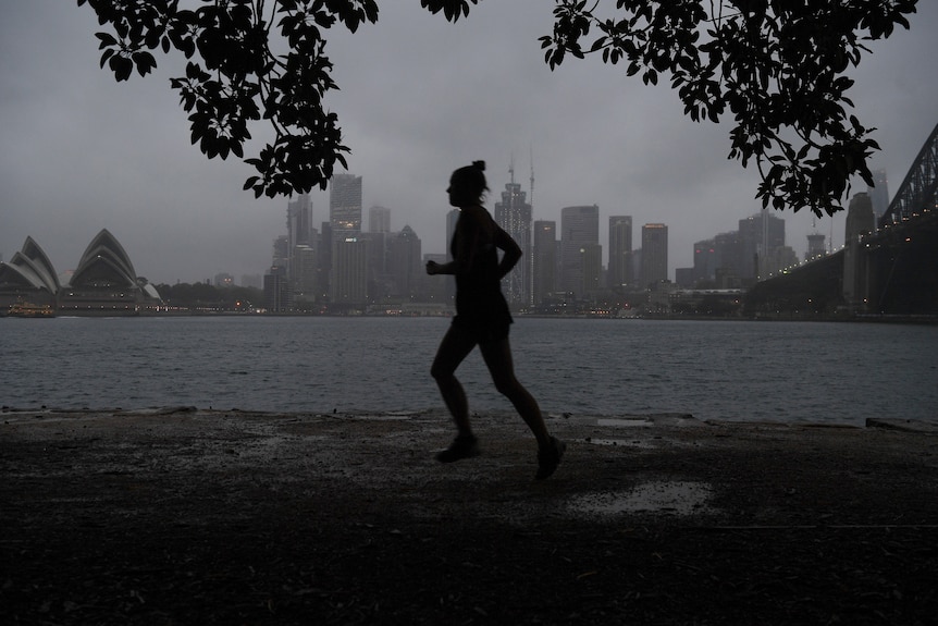 A female jogger is silhouetted as she runs past a backdrop of Sydney's CBD on a gray cloudy morning