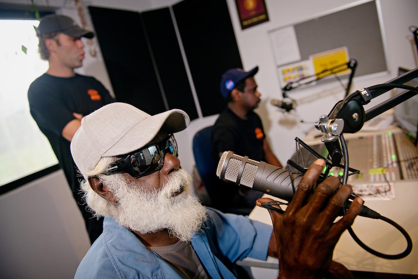 A man wearing sunglasses speaks into a microphone inside a radio studio. 