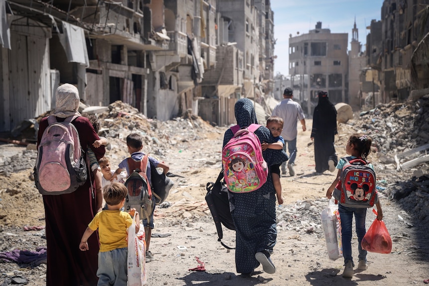 Palestinian women and their children walk through a street littered with rubble and destroyed buildings.
