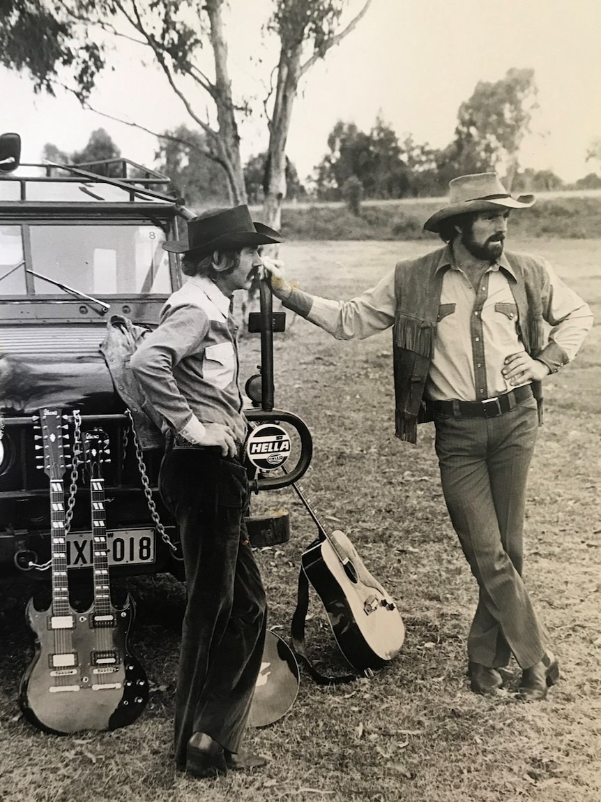 A black and white image of two men in country shirts and wide-brimmed hats standing outside next to a car and guitars.