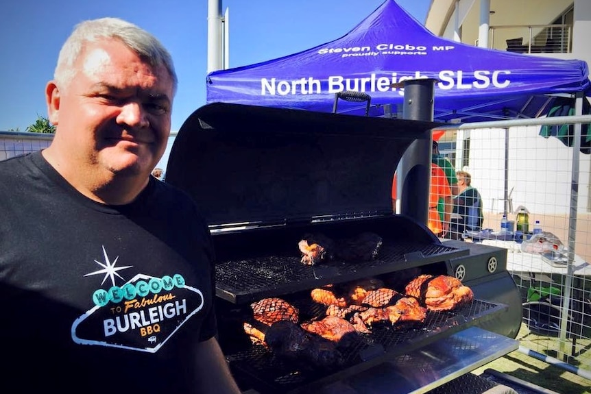 A man stands beside a BBQ near a blue marquee.