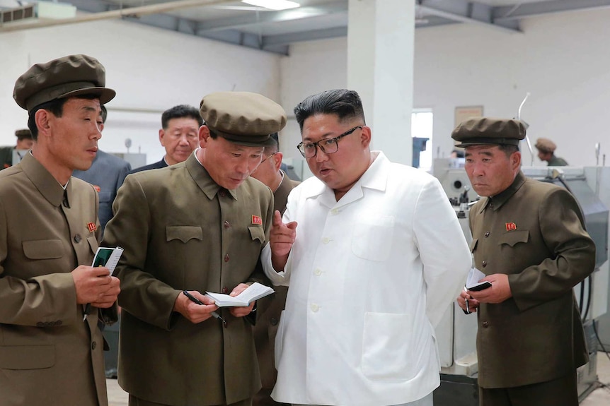 Kim Jong-un has a neutral expression, mid-speech, with his hand up, index finger pointing, as men in miltary garb take notes.