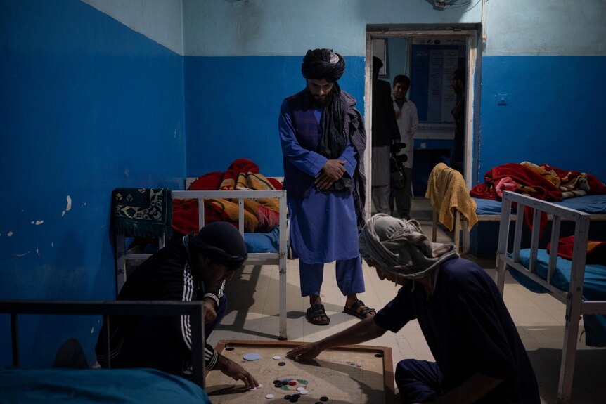 A young Middle Eastern man in traditional dress looks at two men playing a board game on the floor in a shabby dormitory