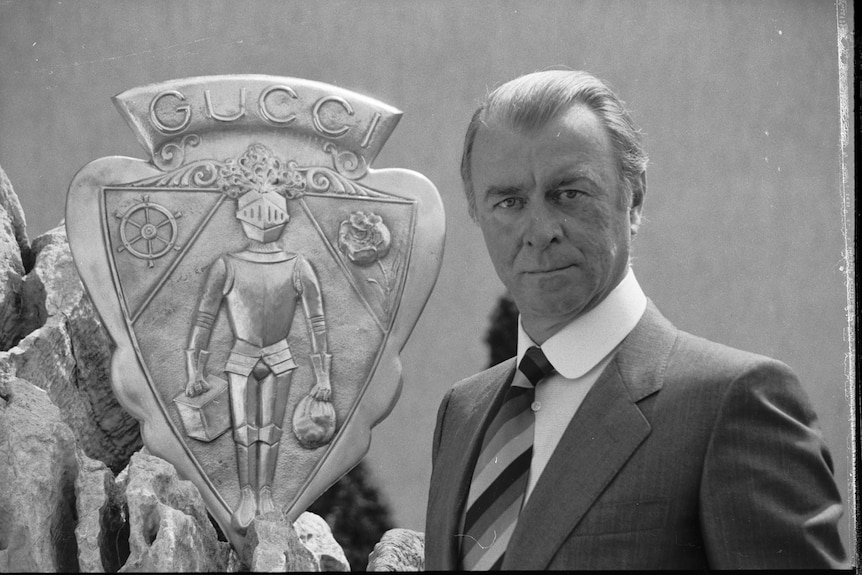 Black-and-white portrait of Roberto Gucci in a sharp suit, standing next to a solid coat of arms bearing the name Gucci