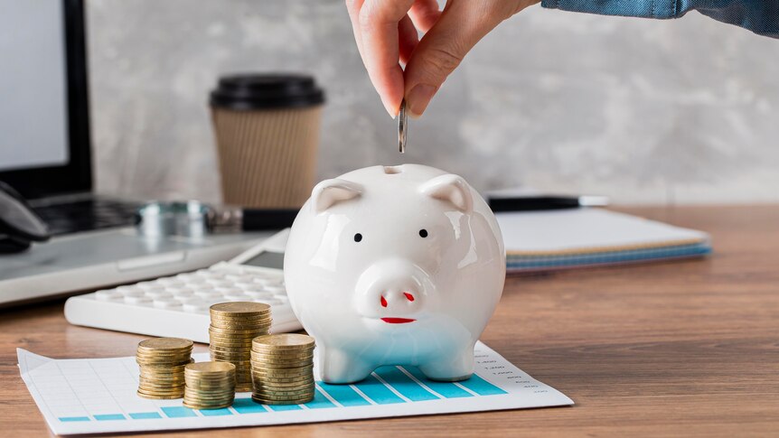 A man about to put a coin into a white piggy bank with other stacks of coins sitting next to the piggy bank