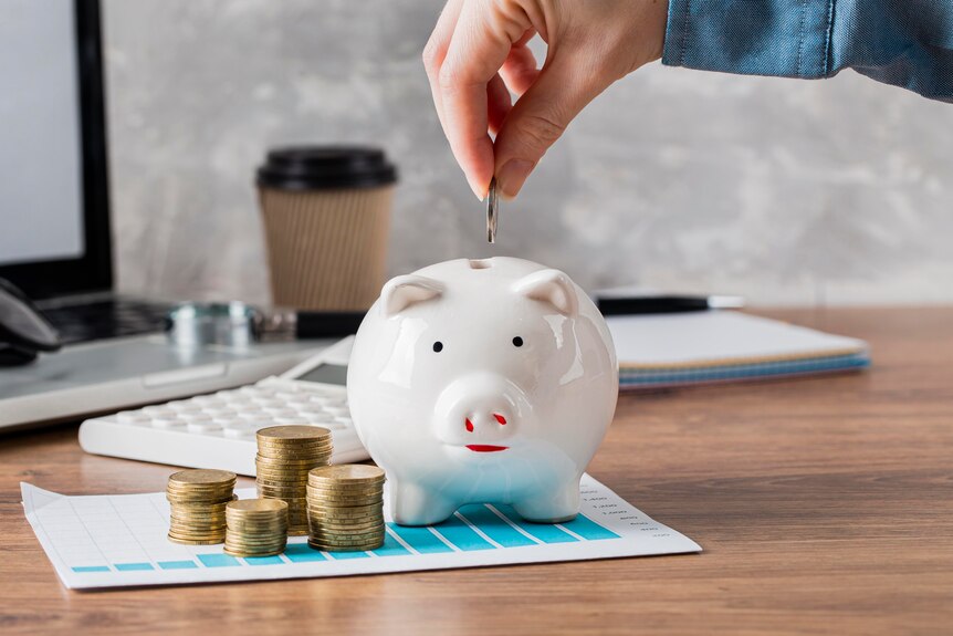 A man about to put a coin into a white piggy bank with other stacks of coins sitting next to the piggy bank