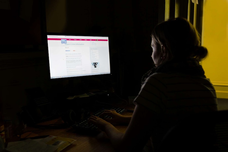 Woman sitting in dark room looking at Crime Stoppers website on computer