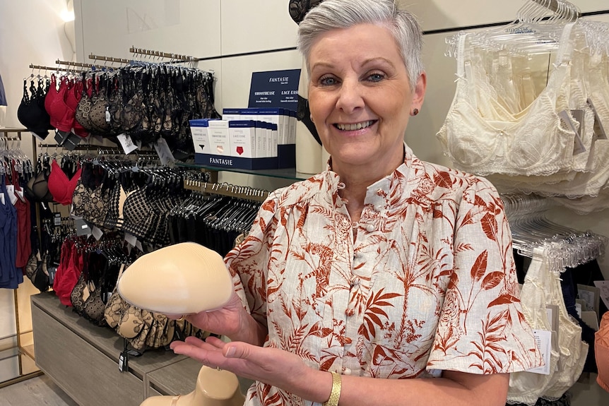 A woman, standing in front of racks of bras, smiles at the camera while holding a breast-like shape in her hand.