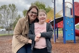 A mother smiles and stands close to her daughter with a playground in the background