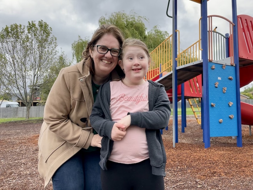 A mother smiles and stands close to her daughter with a playground in the background