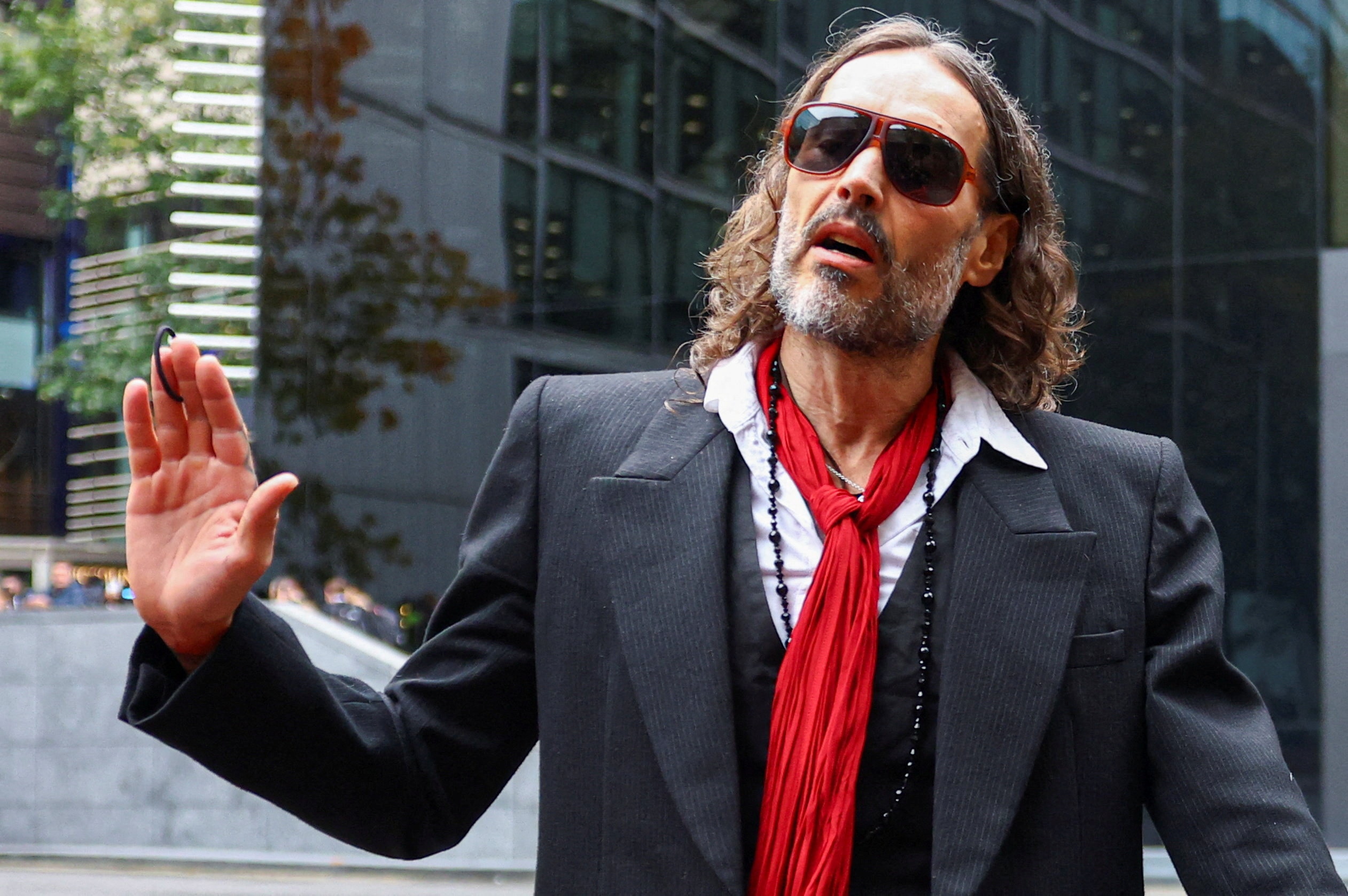 A man, wearing a suit jacket and red scarf and sunglasses, waves