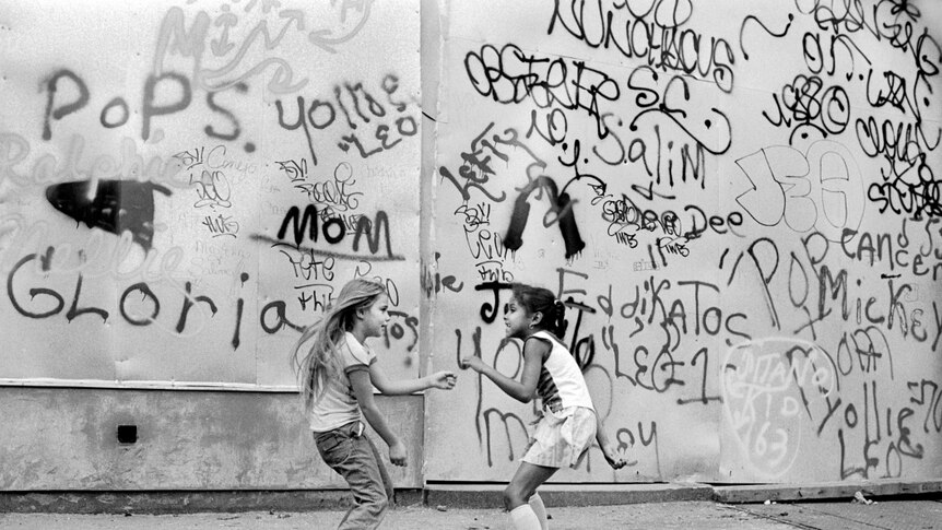 Two young girls playing on street in front of a heavily graffitied wall.