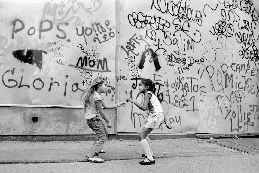 Two young girls playing on street in front of a heavily graffitied wall.