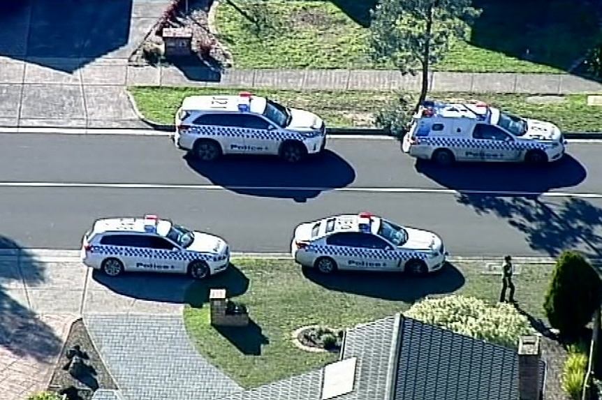 Four police cars parked on nature strips outside a house in a suburban road.