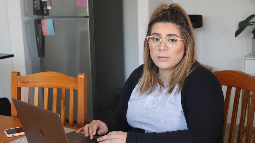 A pregnant woman sits at her living room table searching for jobs on a laptop