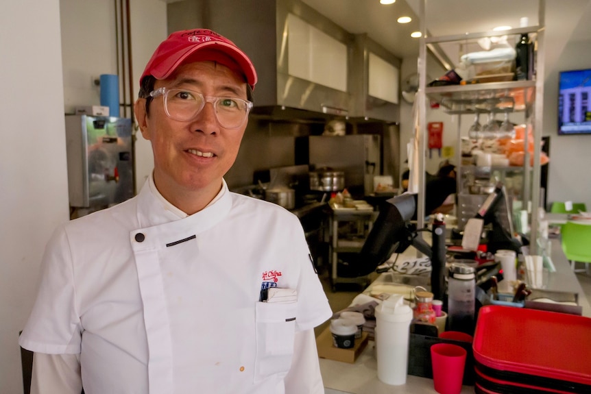 A man wearing a white chef's uniform and a red cap standing in front of a kitchen and cashier space.