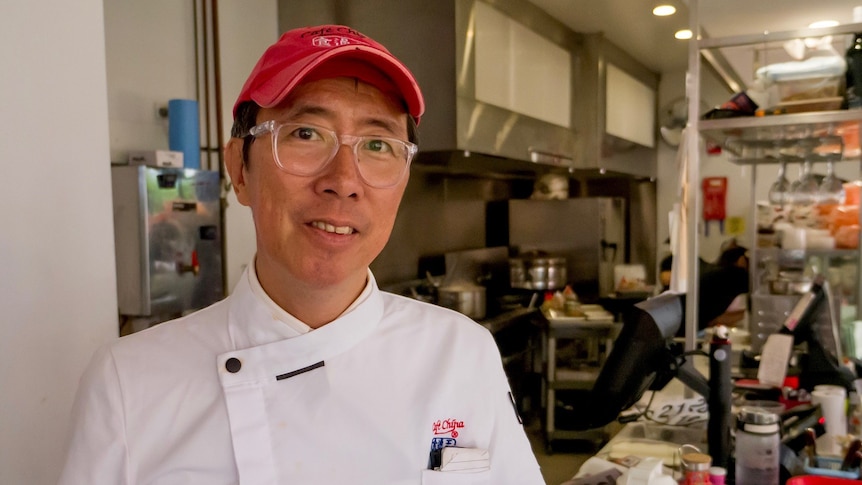 A man wearing a white chef's uniform and a red cap standing in front of a kitchen and cashier space.