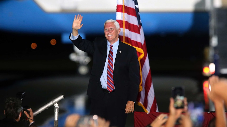 A man in a suit smiles and waves while standing on a stage in front of a US flag and a large aircraft