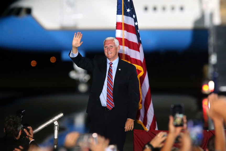 A man in a suit smiles and waves while standing on a stage in front of a US flag and a large aircraft