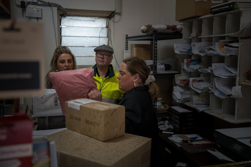 Em, Ash and local postie examine a package behind a desk.