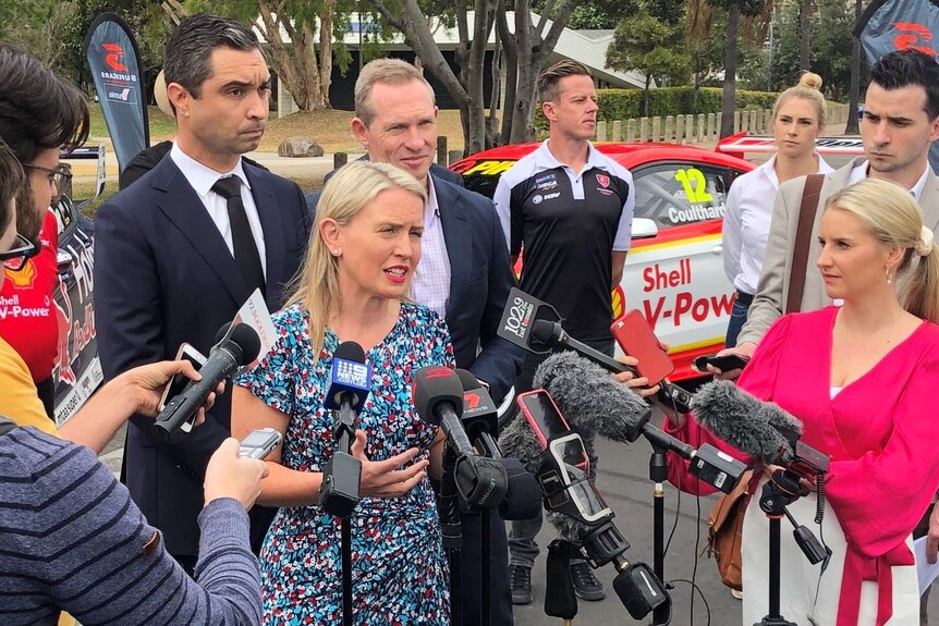 Kate Jones speaks surrounded by media and microphones in front of a V8 car
