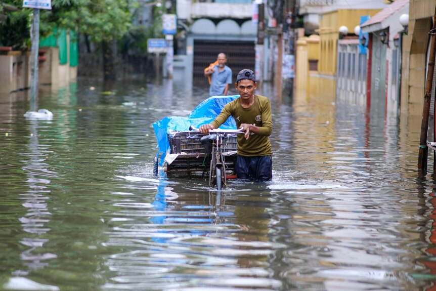 A slim young man with baseball cap pushes his cart through flood waters in south Asian setting