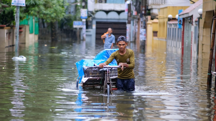 A slim young man with baseball cap pushes his cart through flood waters in south Asian setting