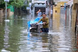 A slim young man with baseball cap pushes his cart through flood waters in south Asian setting