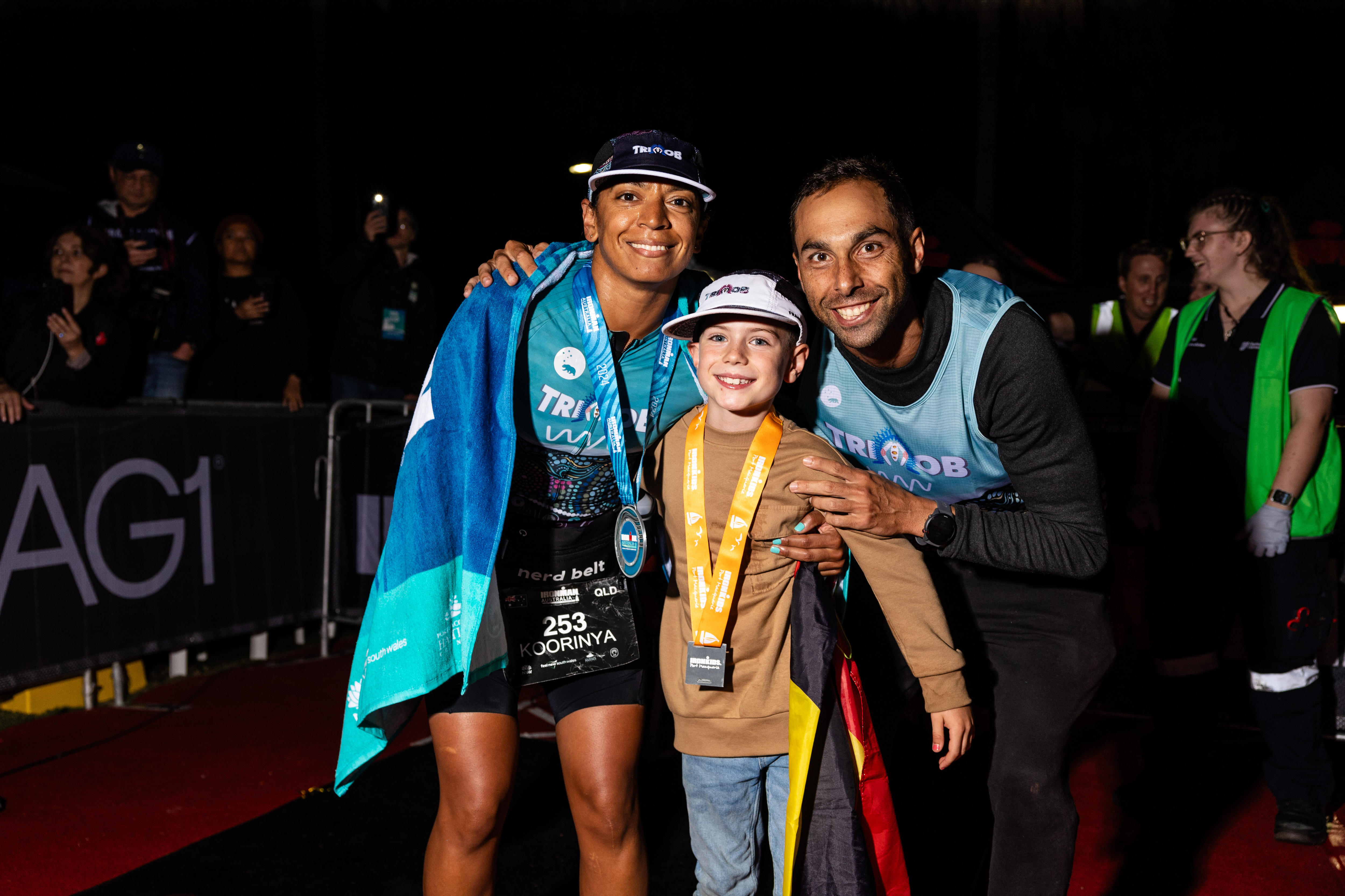 Two Indigenous athletes wearing medals and a small boy hug one another at the finish line of a triathlon.