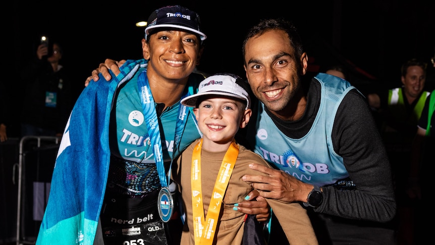 Two Indigenous athletes wearing medals and a small boy hug one another at the finish line of a triathlon.
