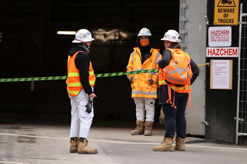 Women in hi-vis orange vests and hard-hats stand outside a workplace with HAZCHEM signs around.