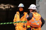 Women in hi-vis orange vests and hard-hats stand outside a workplace with HAZCHEM signs around.