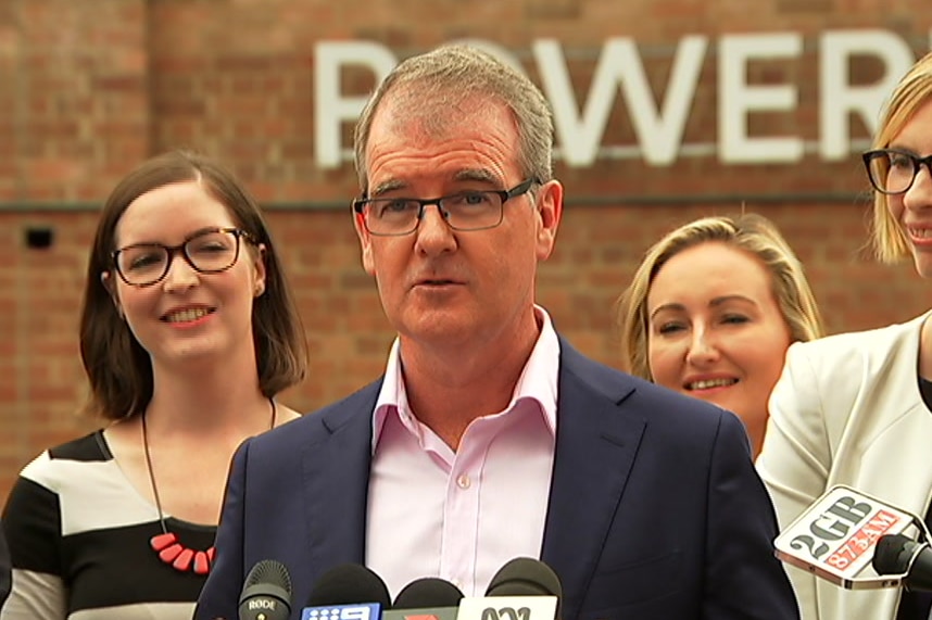 A man in a suit standing in front of political candidates talking to journalists.