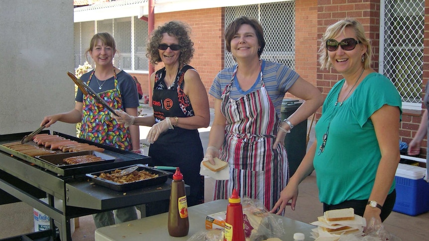 Four women wearing aprons and smiling stand behind a barbecue with sausages and a table with bread and tomato sauce bottles.