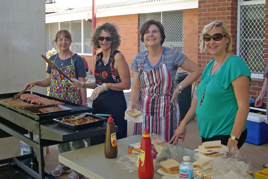 Four women wearing aprons and smiling stand behind a barbecue with sausages and a table with bread and tomato sauce bottles.