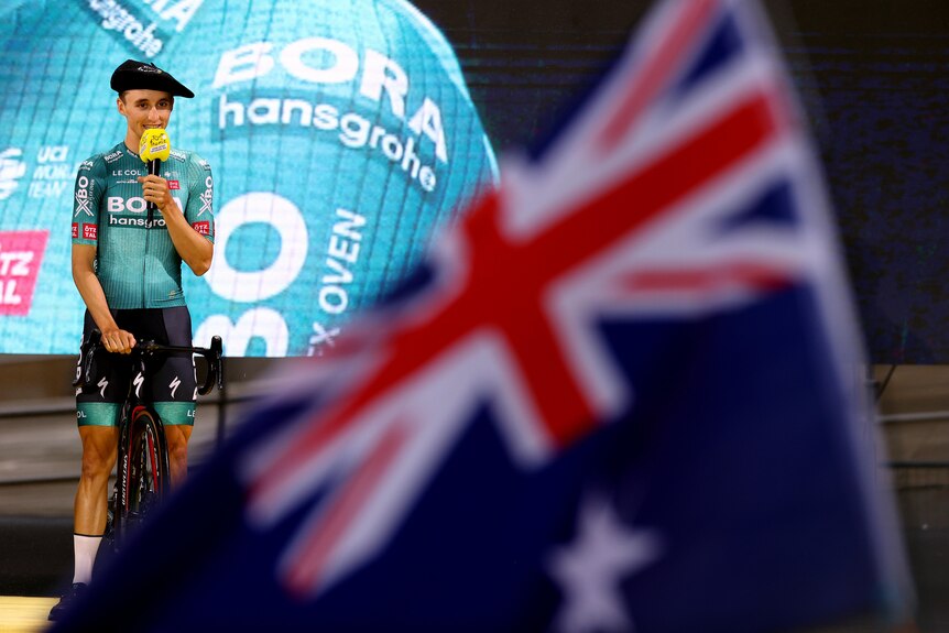 A man speaks on stage while an Australian flag can be seen in the foreground.