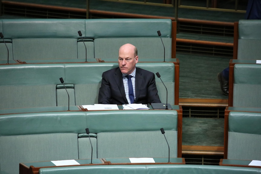 A bald man in a suit sits alone at a row of benches.