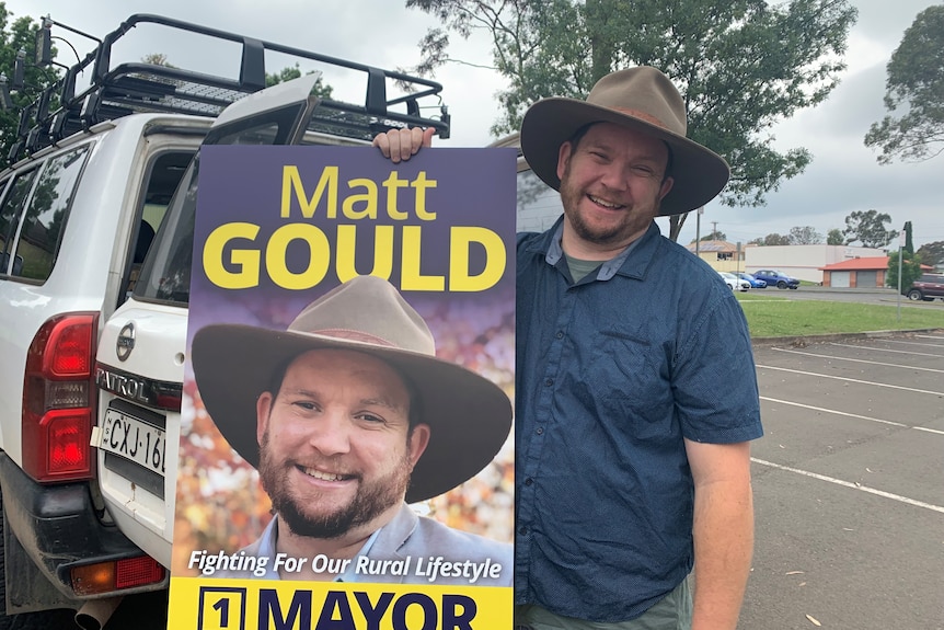 A man wearing a hat stands holding his Mayoral poster
