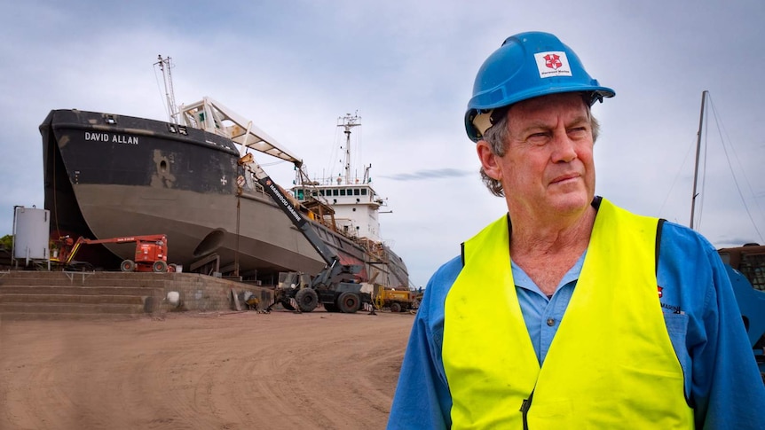 A man in a hard hat with a large ship behind him in a dry dock being repaired