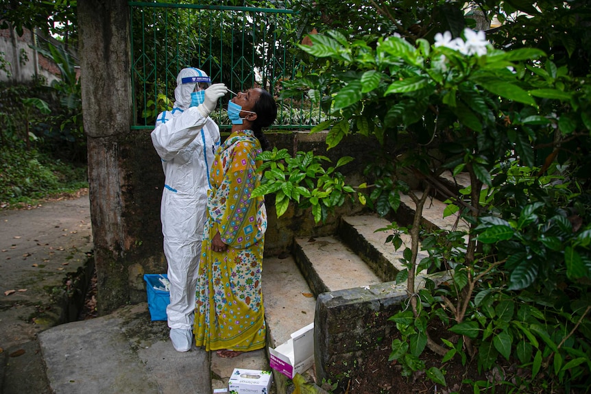 A health worker in PPE uses a nasal swab to test a woman who is holding her head back while standing on steps.