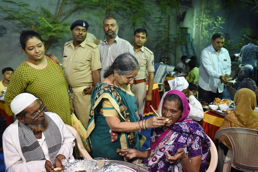 A woman wearing a green sari offers a woman seated before her a piece of fruit while a crowd watches on.