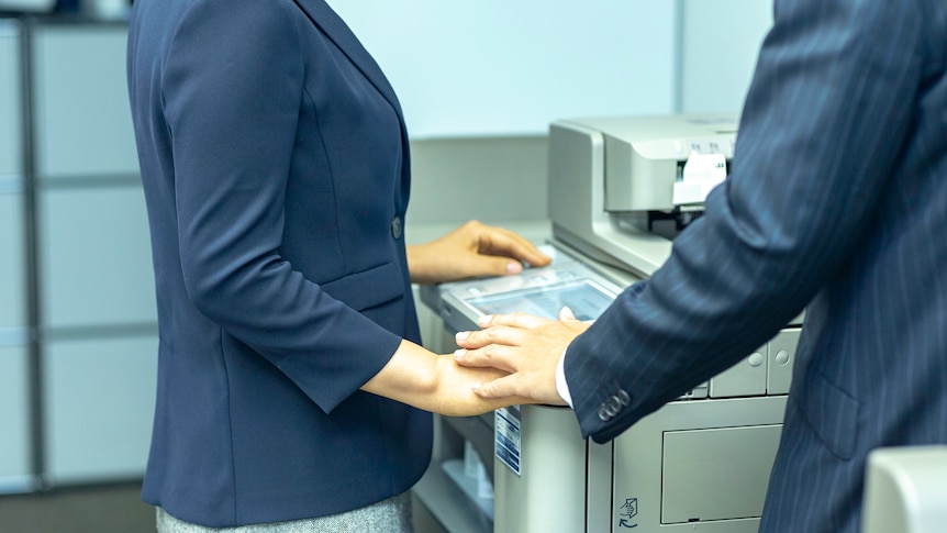 two people hold hands discreetly at a printer in an office.