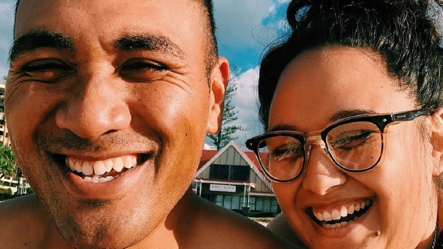 A man and a woman with dark hair and dark skin tones smiling with a blue sky in the background.