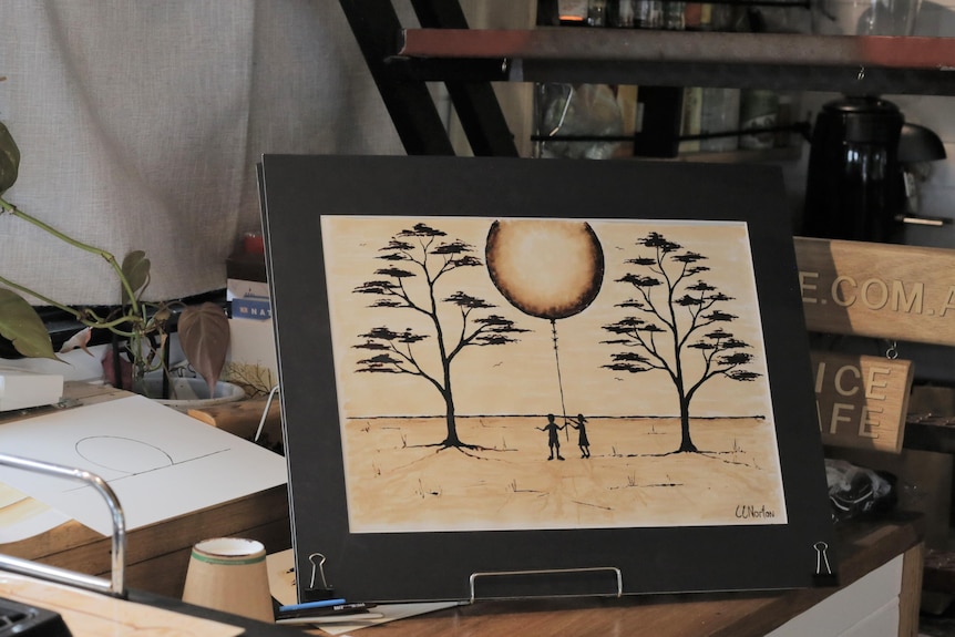 A painting on a small easel on a desk, depicting two trees, two people and a moon. 